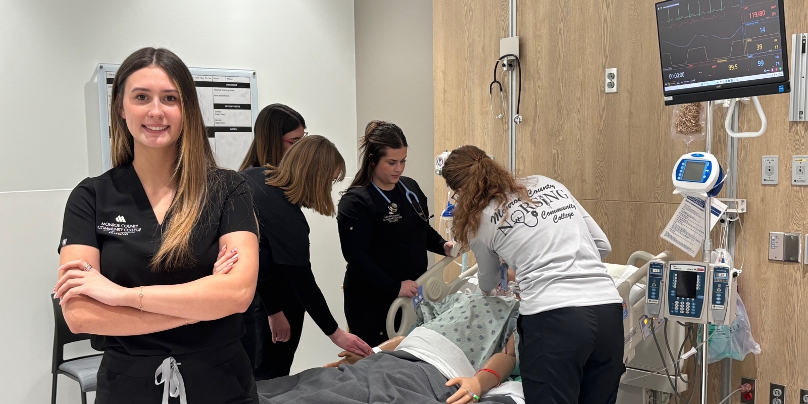 Nursing student posing in lab in front of instructor and other students working on a simulated patient.