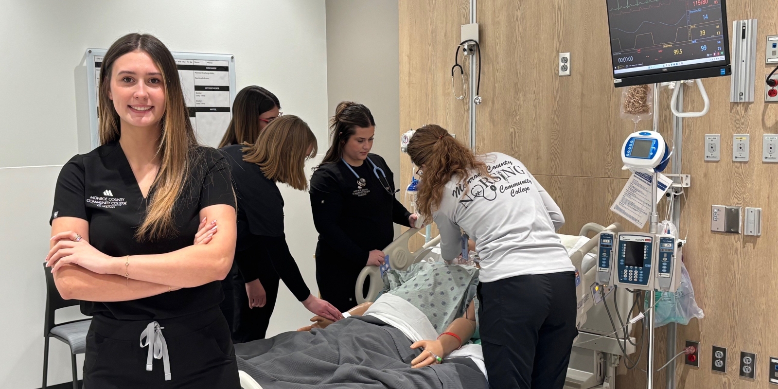 Nursing student posing in lab in front of instructor and other students working on a simulated patient.